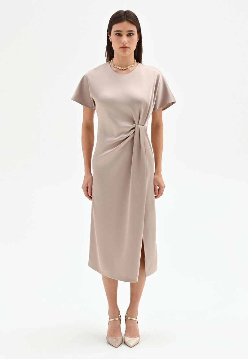 Vrouw staand in een beige midi-jurk met korte mouwen, zijstrikkdetail, split, gouden ketting en gouden enkellaarzen met bandje.