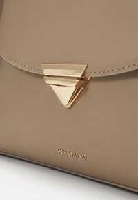Beige konstläderhandväska med ett triangulärt guldmetallspänne och sömdekor. Har varumärket "ANNA FIELD" i präglad text.
