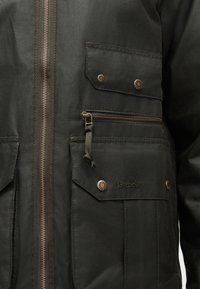 Μπροστινό μέρος σκούρου πράσινου μπουφάν με κάθετο φερμουάρ, τσέπες με καπάκι και ορειχάλκινα κουμπιά τύπου σφικτήρα, οριζόντια τσέπη με φερμουάρ και κεντημένο λογότυπο Barbour.