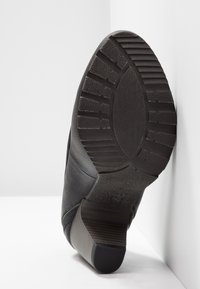 Chaussure en cuir noir avec une semelle texturée et un talon moyen. Présente un design incurvé et des coutures visibles sur les côtés.