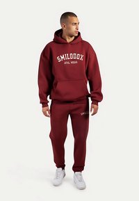 Bordeauxrode hoodie en joggingbroek set, katoenmix, met voorzak, geribbelde boorden en witte logo-tekst. Draagt witte sneakers.