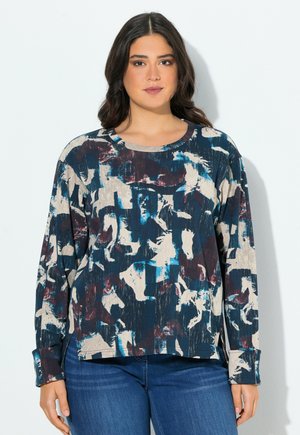 Femme portant un haut à manches longues avec des motifs de chevaux abstraits en bleu, beige et bordeaux, associé à un jean bleu, se tenant contre un mur blanc.