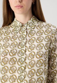 Camicia con bottoni caratterizzata da un motivo geometrico ripetuto in oro su sfondo crema. Tessuto morbido con colletto classico e bottoni a vista.