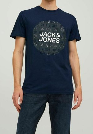 Man wearing navy blue t-shirt with bold "JACK & JONES" text inside a circle, paired with dark blue jeans, standing against plain background.
