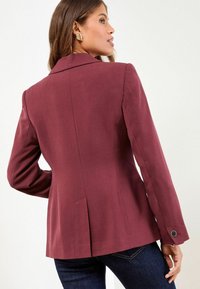 Bordeaux blazer met een gestructureerd ontwerp, enkele achterste split en knoopdetail op de mouwen, gecombineerd met donkere jeans.