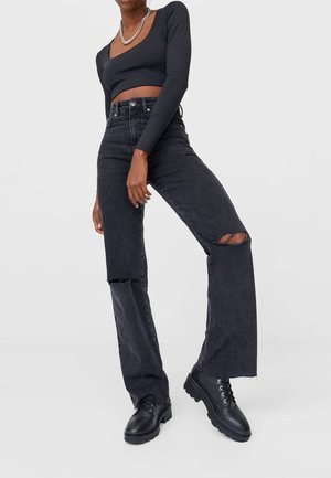 Kobieta ubrana w czarną krótka bluzkę z długim rękawem, czarne, wysokowalne podarte dżinsy, czarne masywne buty sznurowane oraz srebrny łańcuszek na szyi.