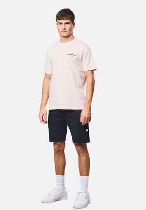 Junger Mann steht, trägt ein hellrosa T-Shirt, schwarze knielange Shorts, weiße Socken und weiße Turnschuhe vor weißem Hintergrund.