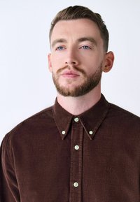 Brązowa koszula z korduroy z kołnierzykiem button-down, z zielonymi guzikami i haftowanym logo po lewej stronie.