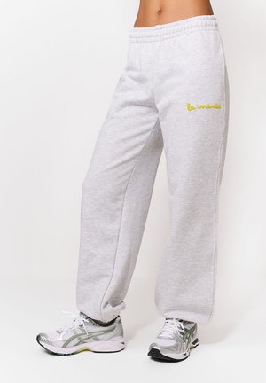 Osoba stojaca na bielom pozadí, oblečená v svetlosivých teplákoch s výšivkou "la maman" v žltej farbe a bielych športových topánkach.