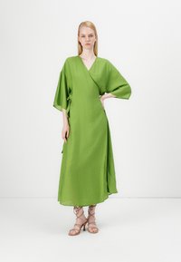 Grønn omslagskjole med vide ermer, laget av teksturert stoff, har en sideknute og midi-lengde, kombinert med strappy sandaler.