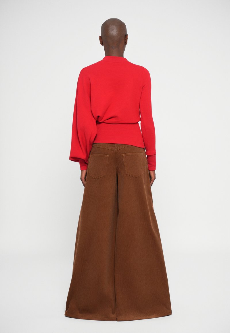 Pull rouge avec de longues manches amples associé à un large pantalon en velours côtelé marron présentant une texture et deux poches arrières.