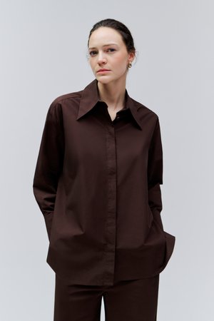 Žena s hnedými vlasmi zviazanými dozadu, nosiaca zlaté kruhové náušnice, voľnú tmavohnedú košeľu na gombíky a zodpovedajúce nohavice na jednofarebnom pozadí.