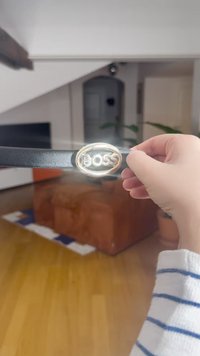 Črni usnjeni pas z ovalno zaponko z zlatim tonom, na kateri je vtisnjen napis "BOSS". Pas drži roka na preprostem notranjem ozadju.