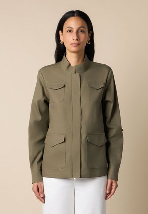 Frau mit langen dunklen Haaren trägt eine olivgrüne Jacke mit vier Klappentaschen und weiße Hose vor einem beigen Hintergrund.