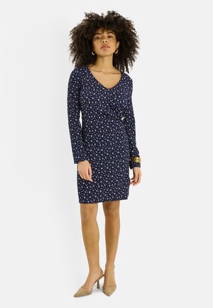 Mujer de pie vistiendo un vestido de manga larga azul marino con pequeño estampado floral blanco y zapatos de tacón beige con punta puntiaguda.