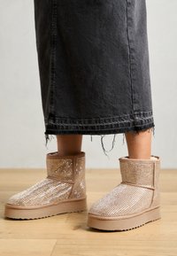 Beige ankelboots dekket med rhinestones, med en avrundet tå og en tykk, teksturert gummisåle, kombinert med et svart denimskjørt.