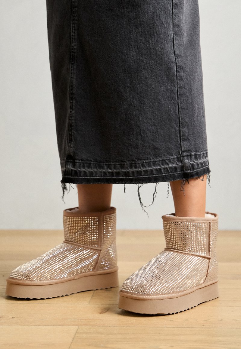 Beige Knöchelstiefel, bedeckt mit Strasssteinen, mit einer runden Zehenpartie und einer dicken, strukturierten Gummisohle, kombiniert mit einem schwarzen Jeansrock.