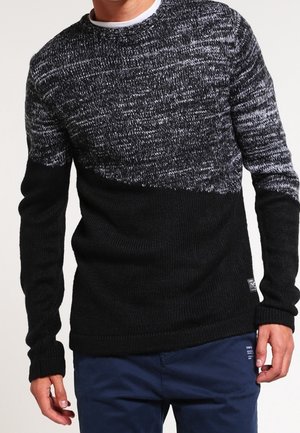 Homme portant un pull en maille bicolore avec la partie inférieure noire et la partie supérieure chinée noir et blanc, associé à un pantalon bleu foncé.