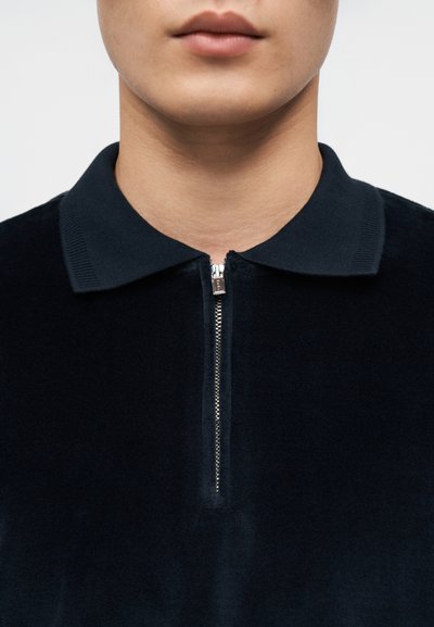 Μπλε ναυτική μπλούζα πόλο με φερμουάρ, υφή υφάσματος, αντίθετο γιακά και κοντό φερμουάρ. Κοντινή θέα που εστιάζει στον σχεδιασμό του λαιμού και του γιακά.
