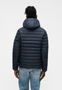 Ναυτικό μπουφάν με ραφές, κουκούλα και εφαρμοστή γραμμή. Συνδυάζεται με ανοιχτόχρωμο μπλε τζιν, παρουσιάζοντας έναν casual και μινιμαλιστικό σχεδιασμό.