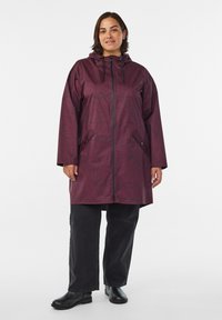 Chaqueta impermeable color marrón burdeos con capucha, que presenta un sutil patrón de hojas, cierre con cremallera y bolsillos laterales. Combinada con vaqueros de pierna ancha negros y botines.