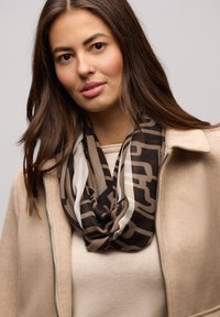 Brun mønstret tørklæde med cremefarvede accenter, båret over en beige sweater og en lys beige jakke. Glat tekstur med et draperet design.