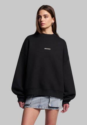 Zwarte oversized sweatshirt met ribbeltextuur en ronde halslijn. Witte tekst "WRSTBHV" op de borst weergegeven. Gecombineerd met een spijkerrok.