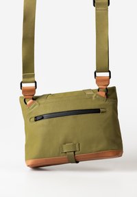 Olivgrüne Umhängetasche mit einem oberen Reißverschluss, braunen Lederakzenten, verstellbarem Schultergurt und einer Vorderreißverschlusstasche. Glatte Textur.