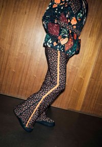 Spodnie dzwony w panterkę z pomarańczowym paskiem po bokach, zestawione z wielopatternowym topem. Tło stanowi drewniana ścianka.