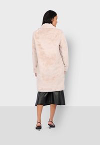 Manteau en fausse fourrure rose clair avec col, manches longues et coupe décontractée, associé à une jupe en cuir noire et des talons avec lanières.
