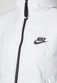 Veste Nike blanche matelassée avec fermeture éclair noire et logo Nike noir sur la poitrine gauche, dotée d'un col montant.