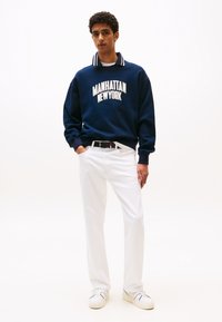 Sweat-shirt bleu marine avec "MANHATTAN NEW YORK" imprimé en blanc, associé à un pantalon blanc et des baskets blanches, avec un col contrastant.