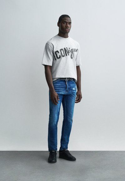 Άνδρας στέκεται μπροστά σε γκρι τοίχο φορώντας γκρι, φαρδιά μπλούζα με το κείμενο «ICONique», στενά μπλε τζιν, μαύρη ζώνη και μαύρα αθλητικά παπούτσια.