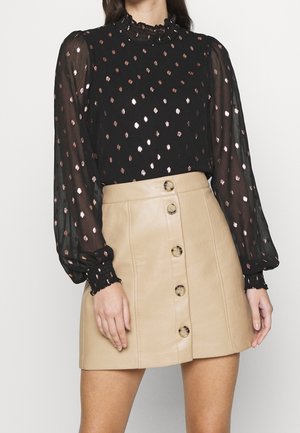 Zwarte blouse met doorschijnende mouwen en een metallic stippenpatroon, gecombineerd met een beige leren minirok met ronde knopen aan de voorkant.