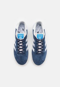 Marineblauwe suède sneakers met een witte driekleurige design, ronde neus, vetersluiting aan de voorkant en een blauwe logo-tag op de tong, met een gestructureerde binnenzool.