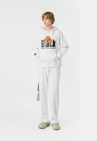 Sudadera blanca con diseño gráfico de perro y texto 'Black', combinada con pantalones blancos y calzado texturizado beige. Diseño simple y moderno.