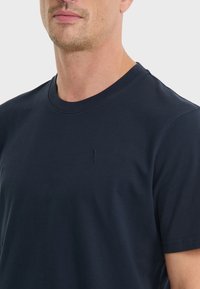 Tummansininen puuvillainen crew neck -t-paita lyhyillä hihoilla, jossa on hienovarainen kirjailtu logo rinnassa. Sileä kangastuntuma.