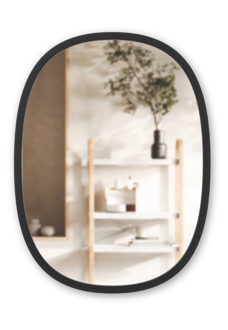 Specchio ovale da parete con una cornice nera, che riflette un ripiano sfocato con accenti di legno chiaro, scatole di stoccaggio bianche e una pianta in un vaso.