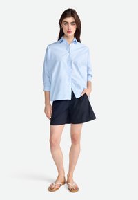 Light blue button-up shirt with long sleeves, paired with dark shorts. Model wears flip-flops; background is plain white.