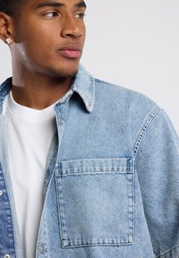 Vaaleansininen farkkupaita, jossa on napitettava kaulus, yksi läpätasku ja näkyvät ompeluyksityiskohdat. Pitää valkoista t-paitaa alla.