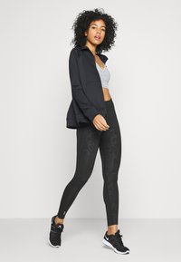 Veste zippée noire sur un crop top gris, associée à des leggings noirs avec un motif léger de peau de serpent et des chaussures de sport noires.