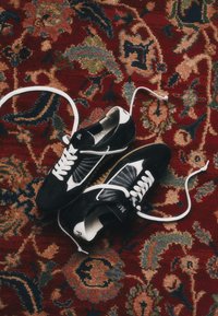 Paire de baskets noir et blanc avec des lacets blancs défaits posée sur un tapis rouge à motifs multicolores.