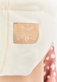 Etykieta z bawełnianej tkaniny na jasnobeżowym tle, z białym tekstem i grafiką oka. Tekst zawiera motywacyjny zwrot.