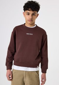 Sudadera marrón de manga larga y cuello redondo, con texto blanco en el pecho. Lleva puesta una camisa blanca y pantalones de pana beige.