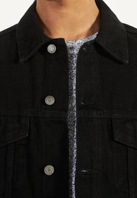 Svart jeansjacka med nedfällbar krage, silverfärgade knappar och framfickor. Under jackan finns en gråmönstrad skjorta.