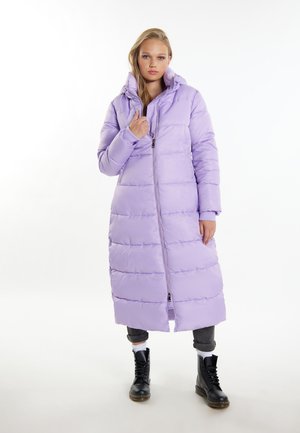 Giovane donna che indossa un lungo piumino lilla chiaro con cappuccio, stivali neri e pantaloni scuri risvoltati su sfondo bianco.
