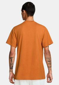 T-shirt a maniche corte in cotone arancio bruciato, con scollo a girocollo e vestibilità comoda, con tatuaggi visibili sulle braccia.