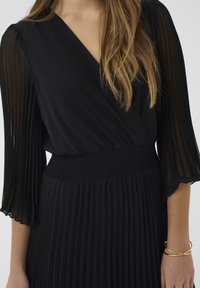 Čierne šaty s plisovanou sukňou, priliehavým pásom s jemným naťahovaním a priehľadnými plisovanými dlhými rukávmi. Mäkčená tkanina elegantne splýva.