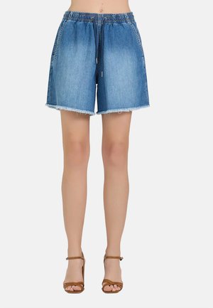 Donna che indossa pantaloncini di jeans blu di media lunghezza con orlo sfrangiato e sandali marroni con tacco alto, in piedi davanti a uno sfondo semplice.