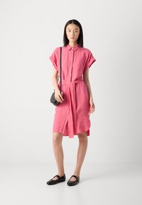 Abito camicia in lino rosa con maniche corte, apertura con bottoni e vita legata, abbinato a scarpe flat nere e una piccola borsa a tracolla.